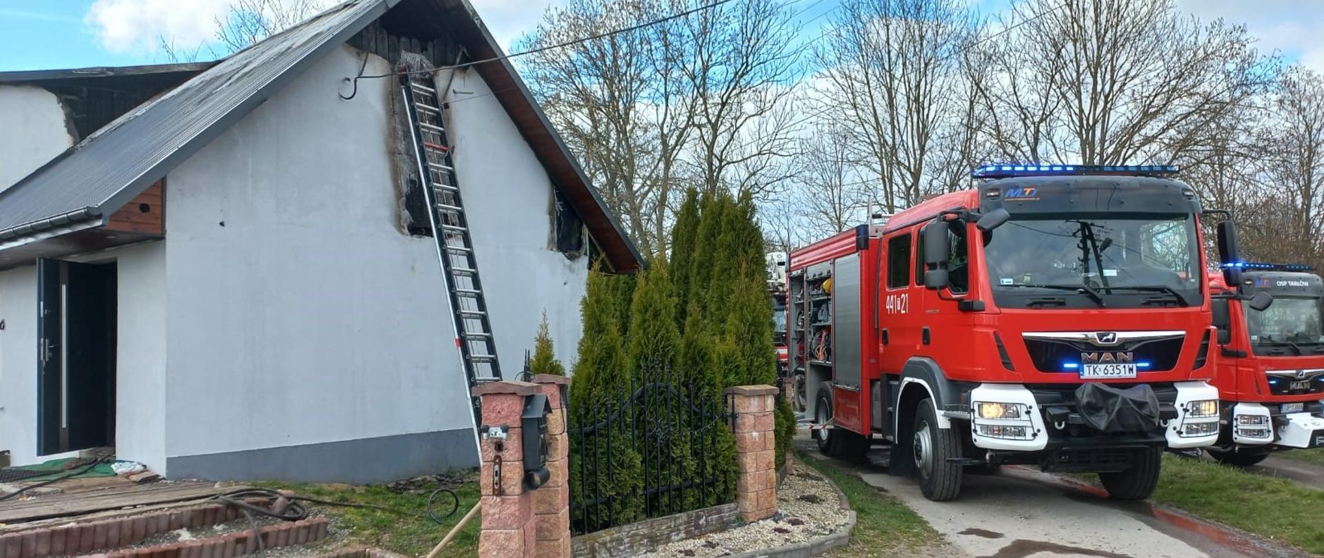 Zdjęcie przedstawia budynek mieszkalny jednorodzinny i ustawioną przy nim drabinę pożarniczą. W tle widać samochody pożarnicze.