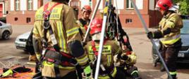 Czterech strażaków ćwiczy wykorzystanie trójnogu ratowniczego do działań wysokościowych w zakresie podstawowym. Trzech strażaków zabezpiecza trójnóg kiedy jeden strażak w uprzęży opuszcza się do studzienki.