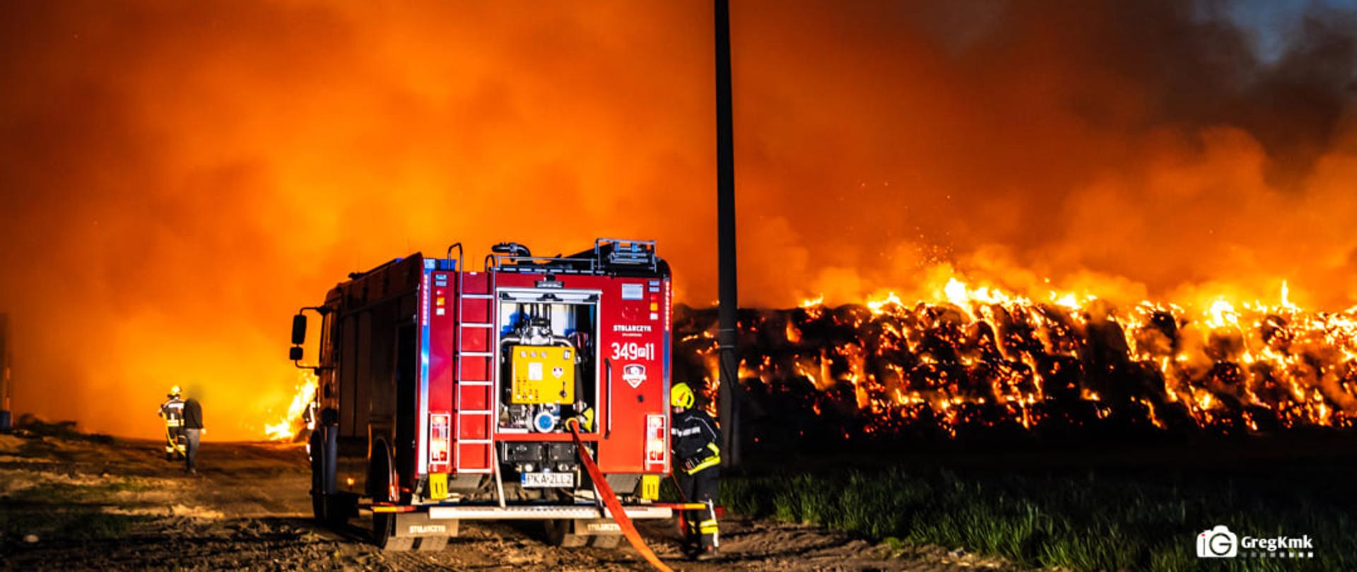 Na zdjęciu widać samochód gaśniczy. Samochód jest w kolorze czerwonym. Posiada na boku wypisane numery operacyjne oraz na dachu światła ostrzegawcze. Za samochodem widać palący się stóg. Obok pojazdu stoi strażak ubrany w ubranie specjalne i hełm. Zdjęcie wykonane w porze nocnej. Nad stogiem widoczna znaczna ilość dymu.
