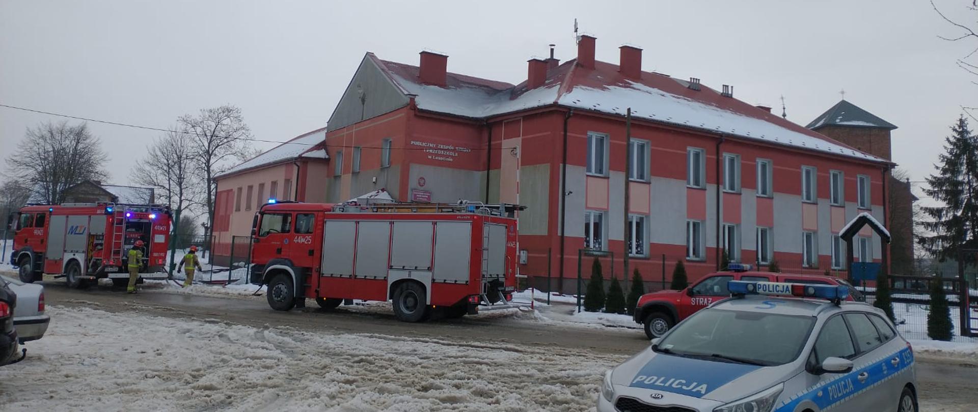 Zdjęcie przedstawia trzy czerwone samochody Jednostek Ochrony Przeciwpożarowej oraz jeden policyjny radiowóz. Samochody stoją na drodze naprzeciwko Publicznej Szkoły Podstawowej w Lasocinie. W tle widać budynek Publicznej Szkoły Podstawowej w Lasocinie.