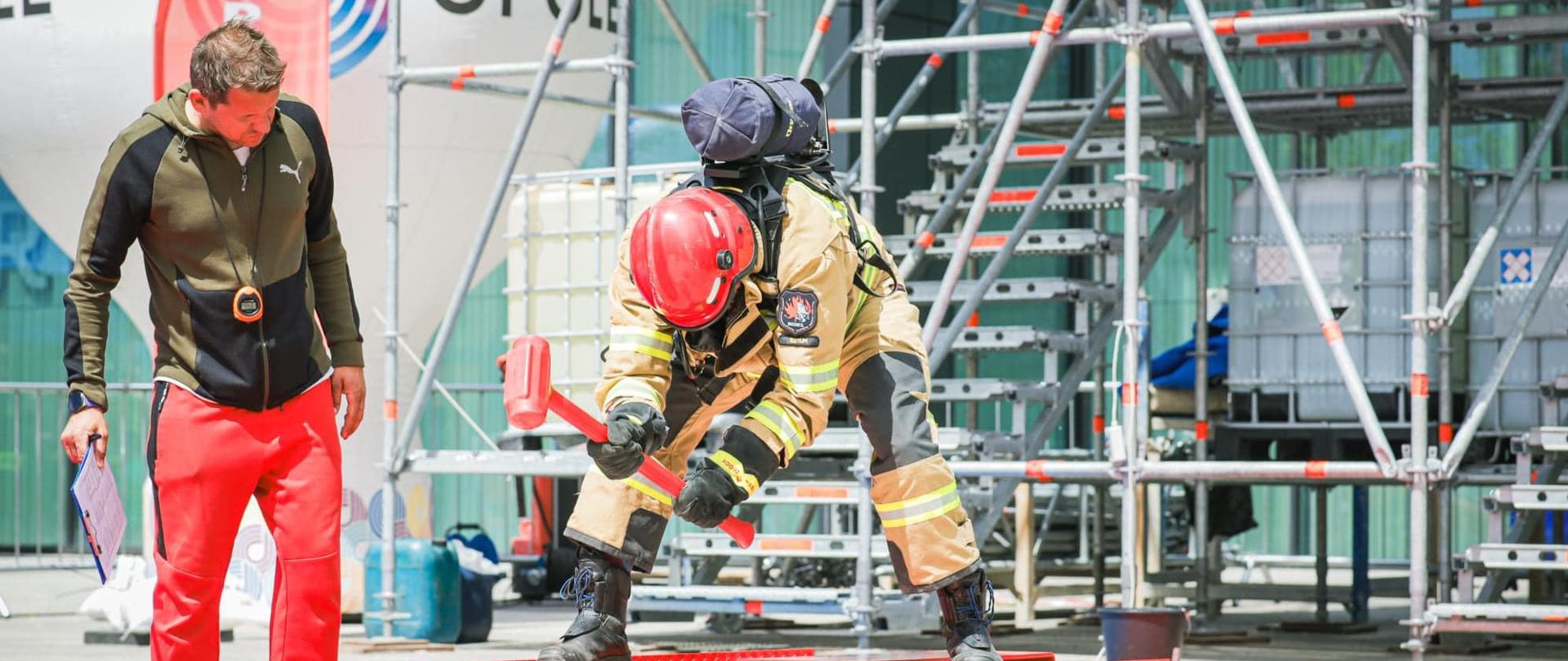 na metalowej konstrukcji w wykroku stoi strażak uderzający młotem o metalową belkę, obok arbitrażuje mu sędzia.