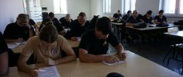 Egzamin teoretyczny kończący szkolenie podstawowe OSP. Strażacy siedzący przy ławkach i rozwiązujących test wiedzy. 