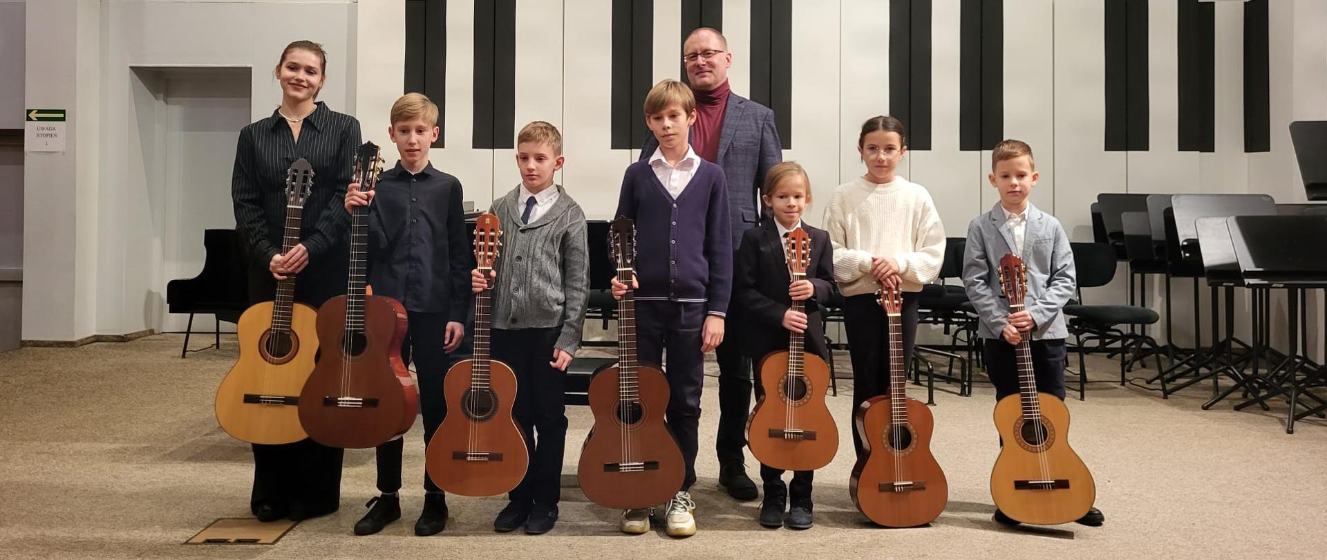 Pięciu chłopców, jedna dziewczynka i nastolatka stoją w jednym rzędzie na estradzie auli, wszyscy trzymają przed sobą gitary. Za nimi stoi mężczyzna.