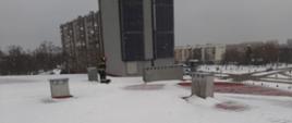 Na pierwszym planie odśnieżany dach KP PSP w Ostrowcu Św. w tle pracujący strażak z łopatą.