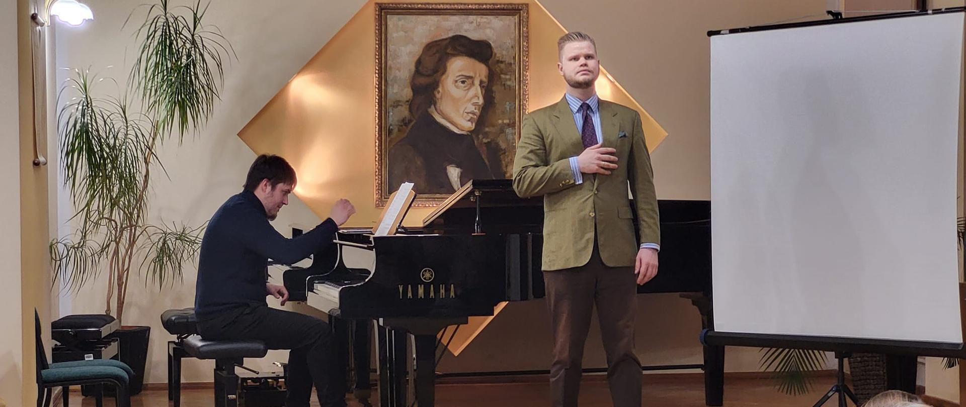 Na zdjęciu od lewej Michał Michalski grający na fortepianie, a po prawej stronie śpiewający David Stoklosa , w tle zdjęcia widoczny portret F. Chopina, na przodzie zdjęciu widoczna publiczność. Zdjęcie wykonane w auli PSM Kłodzko, podczas koncertu z okazji obchodów dnia patrona szkoły - 01 marca 2023 r.