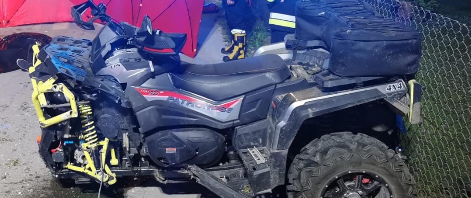 Zdjęcie przedstawia na pierwszym planie uszkodzony pojazd czterokołowy (quad). Na drugim planie widać strażaków w umundurowaniu specjalnym, parawan ochronny oraz karetkę pogotowia z włączoną sygnalizacją świetlną. 