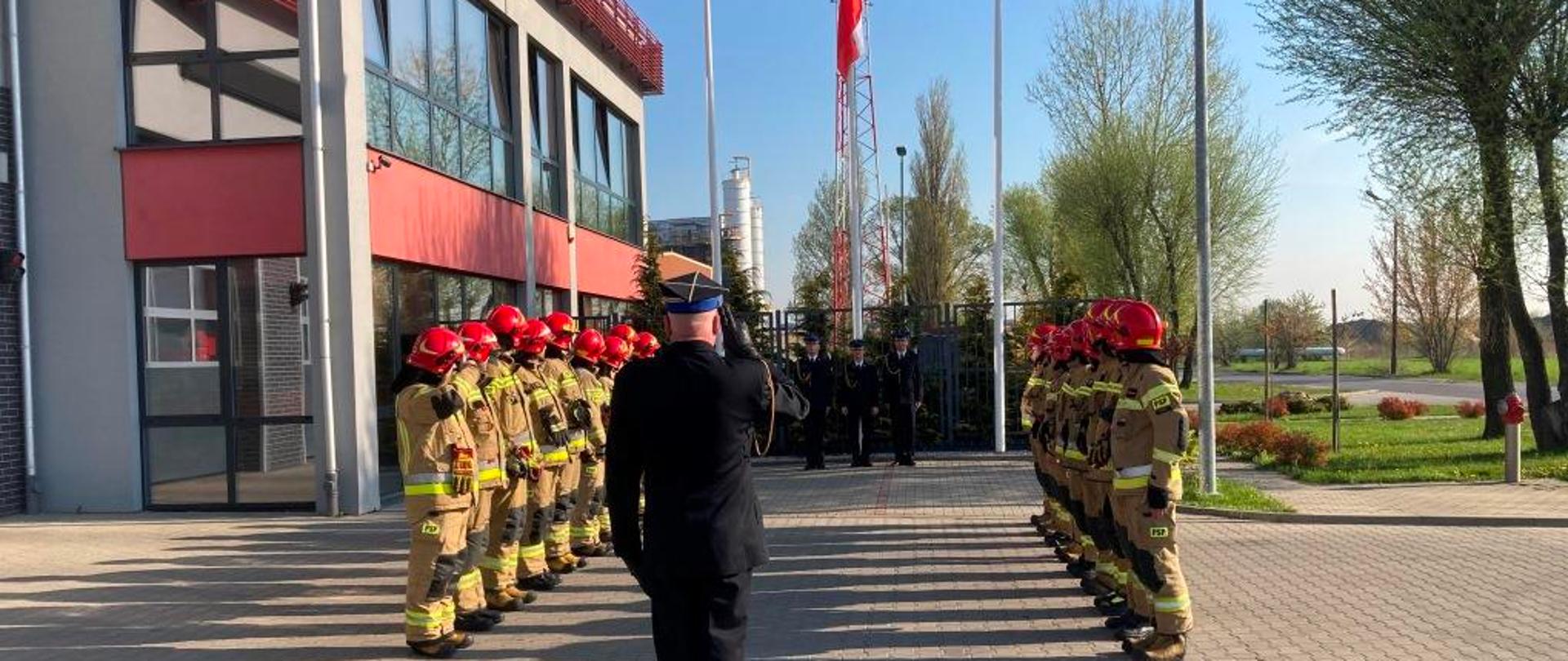 Zdjęcie przedstawia funkcjonariuszy Państwowej Straży Pożarnej. Funkcjonariusze ustawieni w dwóch szeregach zwróconych frontem ku sobie. Ubrani są w ubrania specjalne w kolorze piaskowym. Na głowach pozakładane mają hełmy w kolorze czerwonym. Pomiędzy nimi stoi dowódca jednostki. Ubrany jest w galowy mundur. Strażacy ustawieni są w pozycji zasadniczej „na baczność”podczas podniesienia flagi państwowej na maszt. W oddali widać poczet flagowy, który dokonuje podniesienia flagi. Zdjęcie wykonane przed Jednostką Ratowniczo – Gaśniczą numer 2 w Radomiu. 