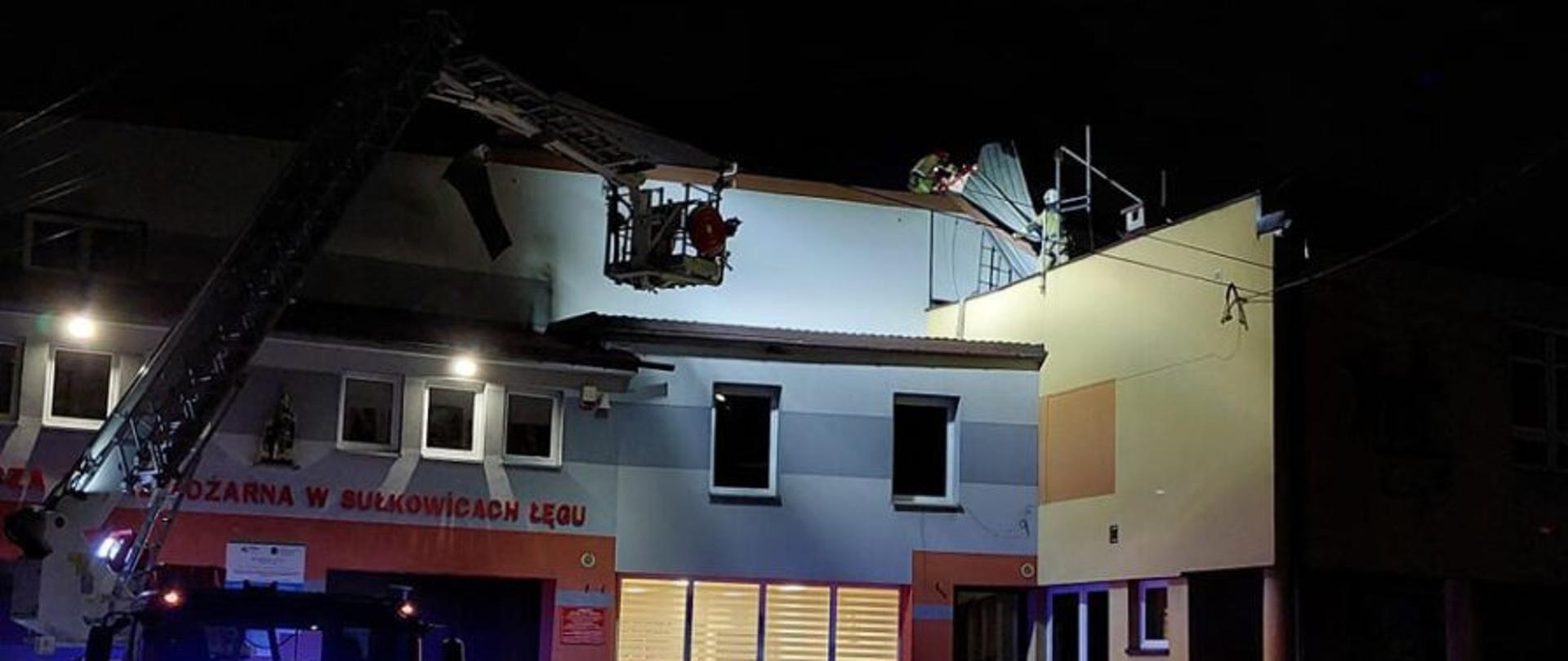 Uszkodzone poszycie dachu na budynku OSP Sułkowice Łęg. Przed remizą podnośnik strażacki i strażacy, którzy będą ściągać uszkodzone elementy dachu. Pora nocna.