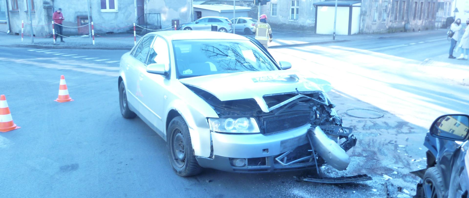 Zdjęcie przedstawia srebrny samochód osobowy typu sedan po zderzeniu z innym autem, który stoi na środku skrzyzowania. W tle widać pachołki ostrzegawcze oraz budynki. 