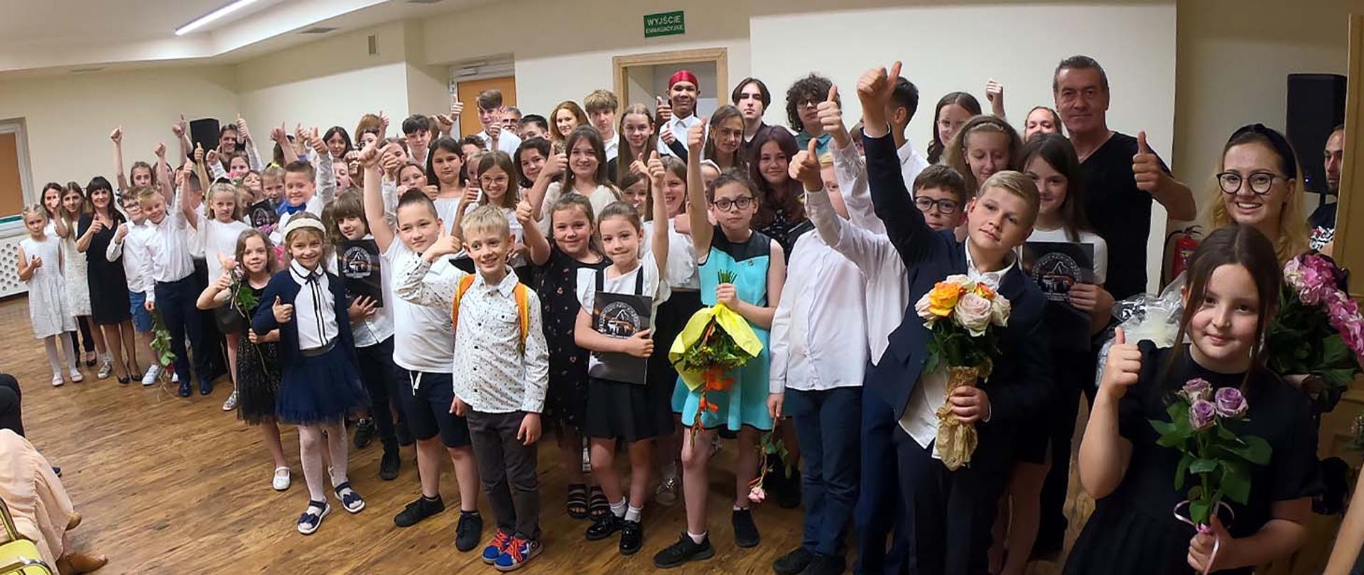 Grupa kilkudziesięciu osób, uczniów i nauczycieli szkoły pozują do zdjęcia w auli szkolnej, wszyscy trzymają w górze uniesione kciuki, niektórzy mają w rękach bukiety kwiatów.