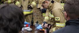 Kilku strażaków w umundurowaniu PSP pochyla się nad stołem, na którym rozłożono sprzęt pomiarowy, narzędzia i próbki wykorzystywane podczas ćwiczeń. Jeden z uczestników trzyma w dłoni urządzenie i pokazuje sposób jego użycia, a pozostali obserwują z bliska. Na drugim planie stoją strażacy OSP oraz wóz strażacki.