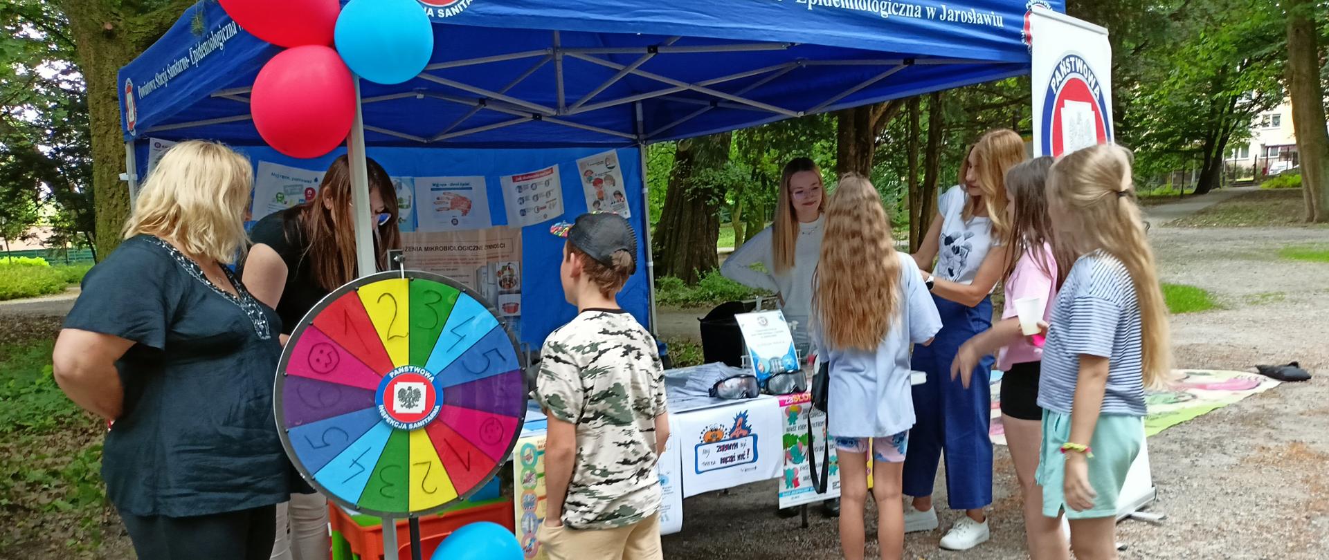 Ludzie gromadzą się wokół stoiska z balonami i kołem do gry w parku.