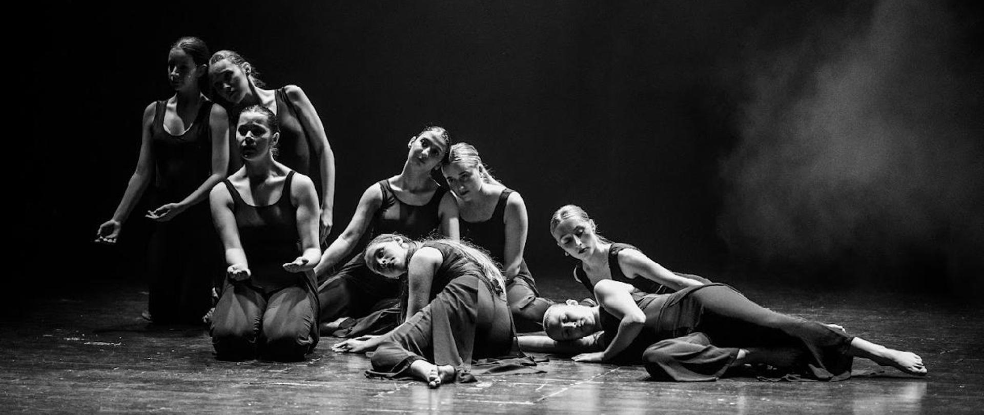 Grupa 8 dziewcząt podczas występu tanecznego. Są w pozach leżących i pół leżących. Każda z nich jest ubrana w czarny kostium na ramiączkach. Znajdują się na scenie. Pada nich delikatne białe światło sceniczne.