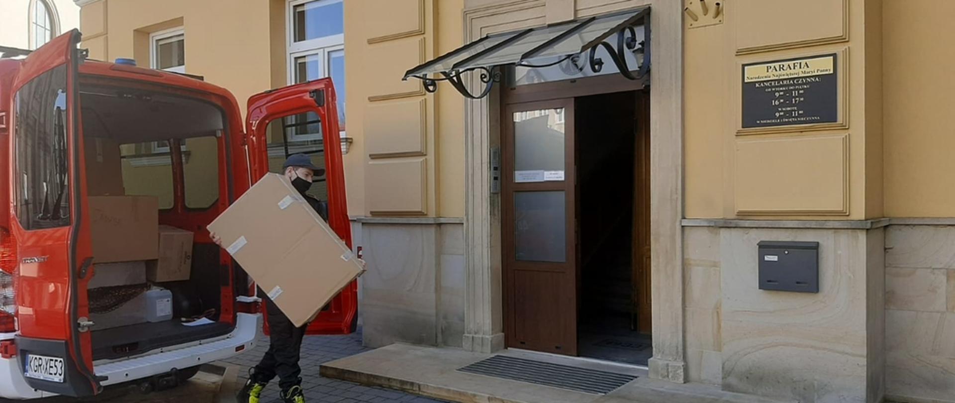 Na zdjęciu widoczny strażak w ubraniu czarnym koszarowym trzymający w rękach dwa kartony z maseczkami ochronnymi. Za plecami strażak widoczny czerwony samochód pożarniczy a przed nim wejście na plebanię. 