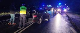 Zderzenie dwóch aut. Droga wojewódzka nr 309 w okolicach Żylic. Na jezdni stoi rozbite audi. Pojazd ma uszkodzoną swoją przednią część. Auto jest koloru ciemnego. Ma uniesioną pokrywę bagażnika. Przed samochodem stoją strażacy i wozy strażackie. Za pojazdem - policjant i kierowca