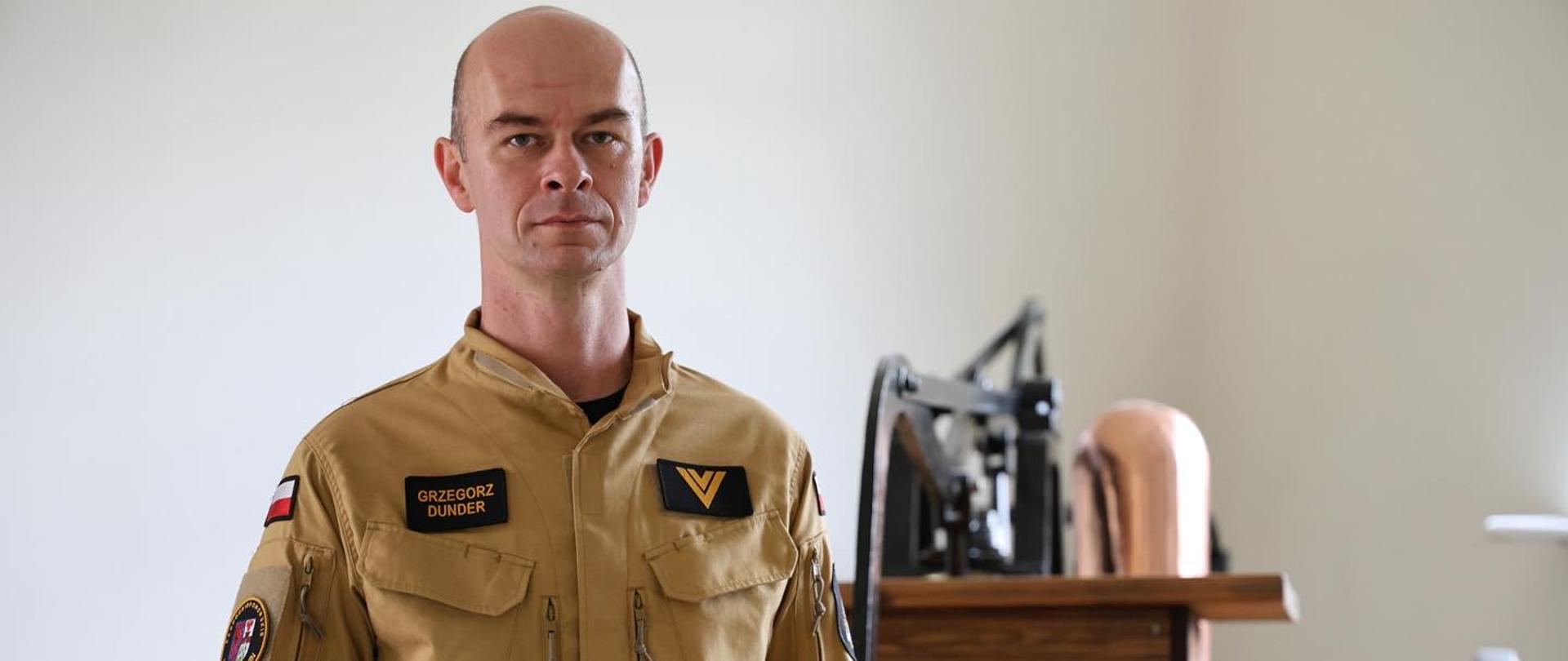 Zdjęcie portretowe, na którym widać strażaka, trzymającego w rękach historyczny hełm. Patrzy prosto w obiektyw z poważnym wyrazem twarzy. Ubrany jest w piaskowy mundur z widocznymi kieszeniami zapinanymi na zamek błyskawiczny. Tło jest jasne, jednolite. Po prawej stronie, za mężczyzną, widać fragment historycznego wyposażenia strażackiego. 