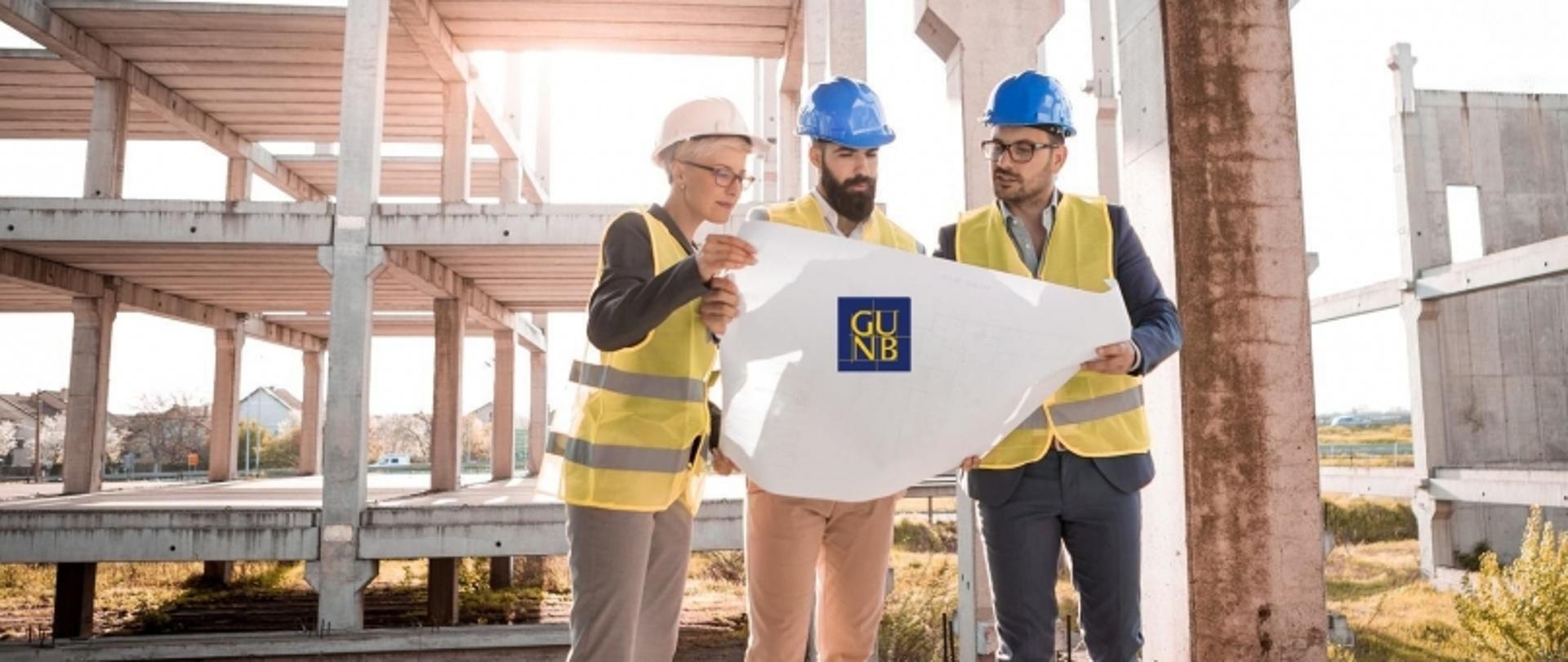 Trzy osoby na placu budowy z planem oznaczonym logo „GUNB”; w tle niedokończony betonowy budynek.