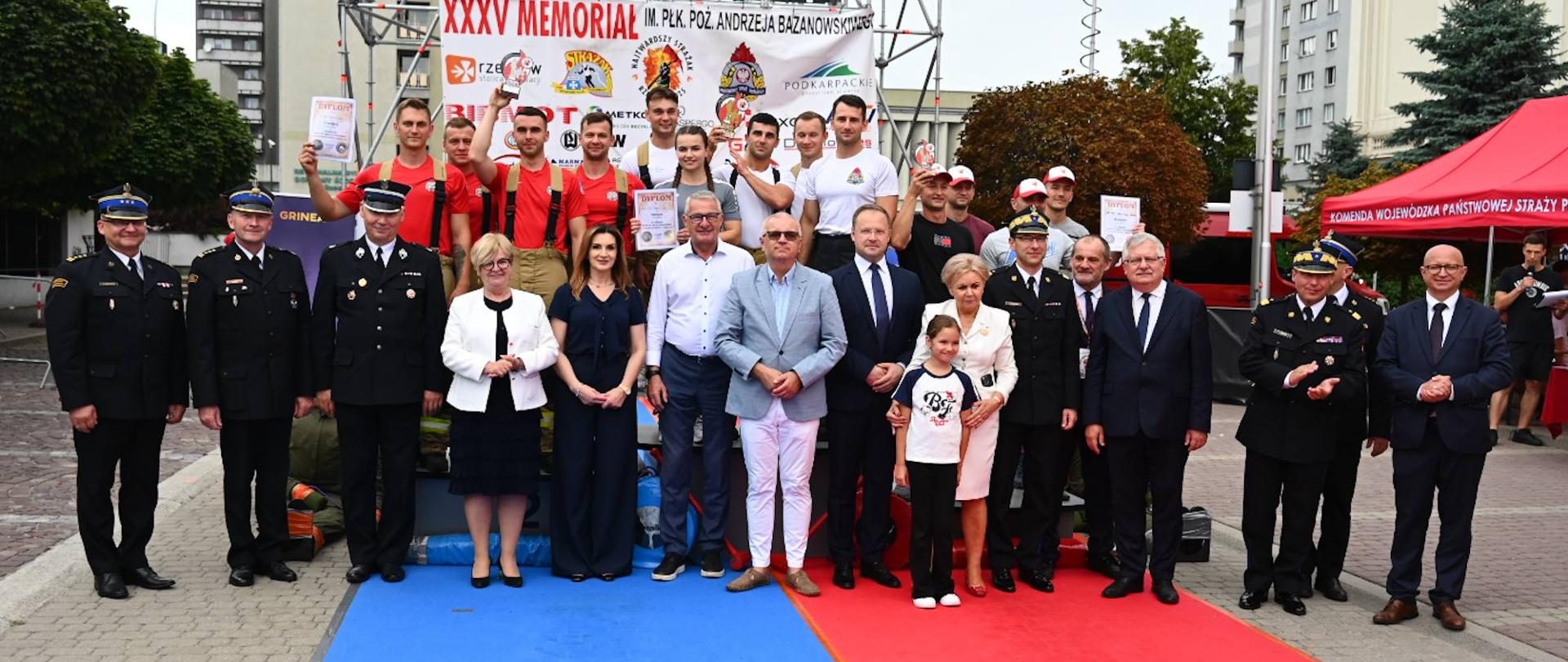 Grupa laureatów i gości honorowych XXXV Memoriału im. płk. poż. Andrzeja Bazanowskiego pozuje wspólnie na podium przed banerem wydarzenia. Strażacy w ubraniach sportowych i galowych stoją obok przedstawicieli władz i organizatorów na tle miejskiej sceny.