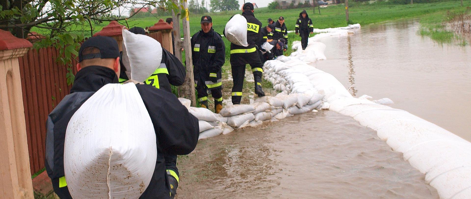 Na zdjęciu strażacy układają worki z piaskiem zabezpieczając pobliską posesję przed zalaniem.