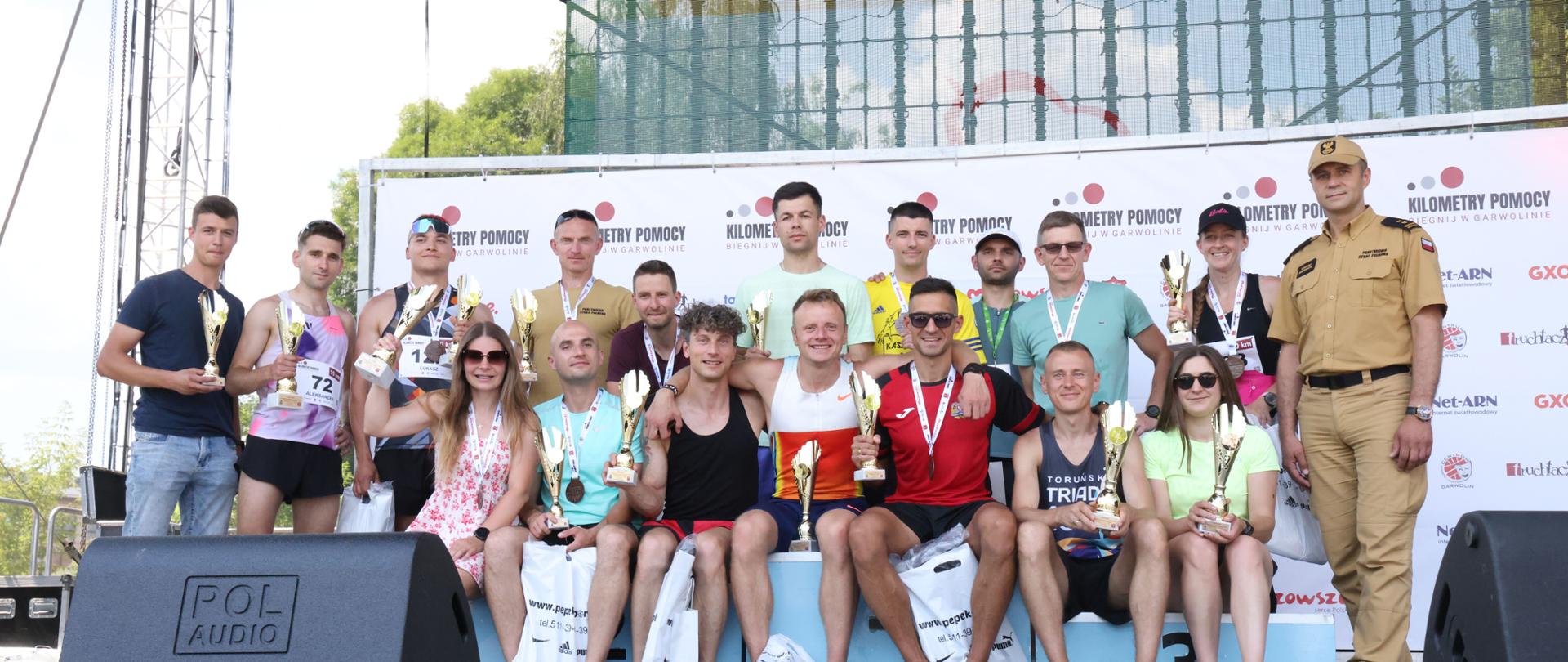 Na scenie zgromadziła się duża grupa uczestników i organizatorów, którzy wspólnie pozują do pamiątkowego, finałowego zdjęcia. Wszyscy są uśmiechnięci, trzymają w rękach zdobyte trofea i wspólnie wypełniają całą przestrzeń wokół podium.