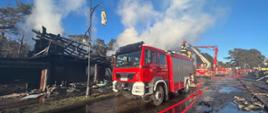 Samochód gaśniczy stoi obok spalonego obiektu, na dachu stoi strażak. Za pojazdem ustawione są kolejne samochody.