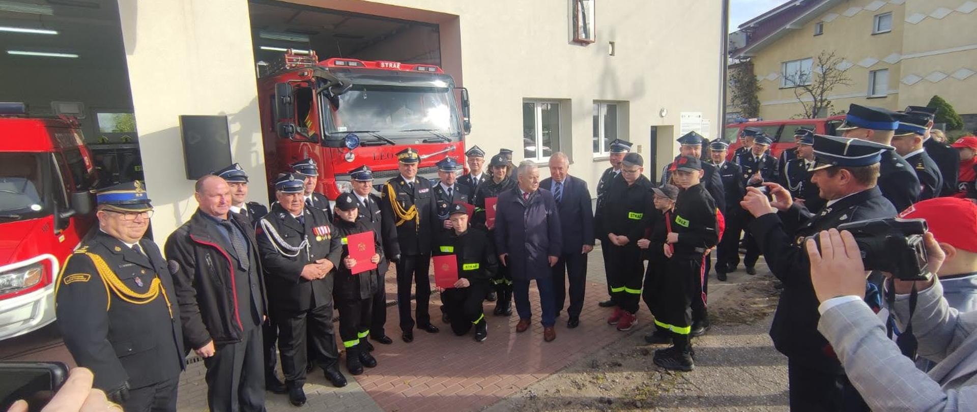 Pomorski komendant wojewódzki Państwowej Straży Pożarnej stoi wspólnie z zaproszonymi gośćmi oraz przedstawicielami Młodzieżowych Drużyn Pożarniczych którzy trzymają czerwone teczki.
