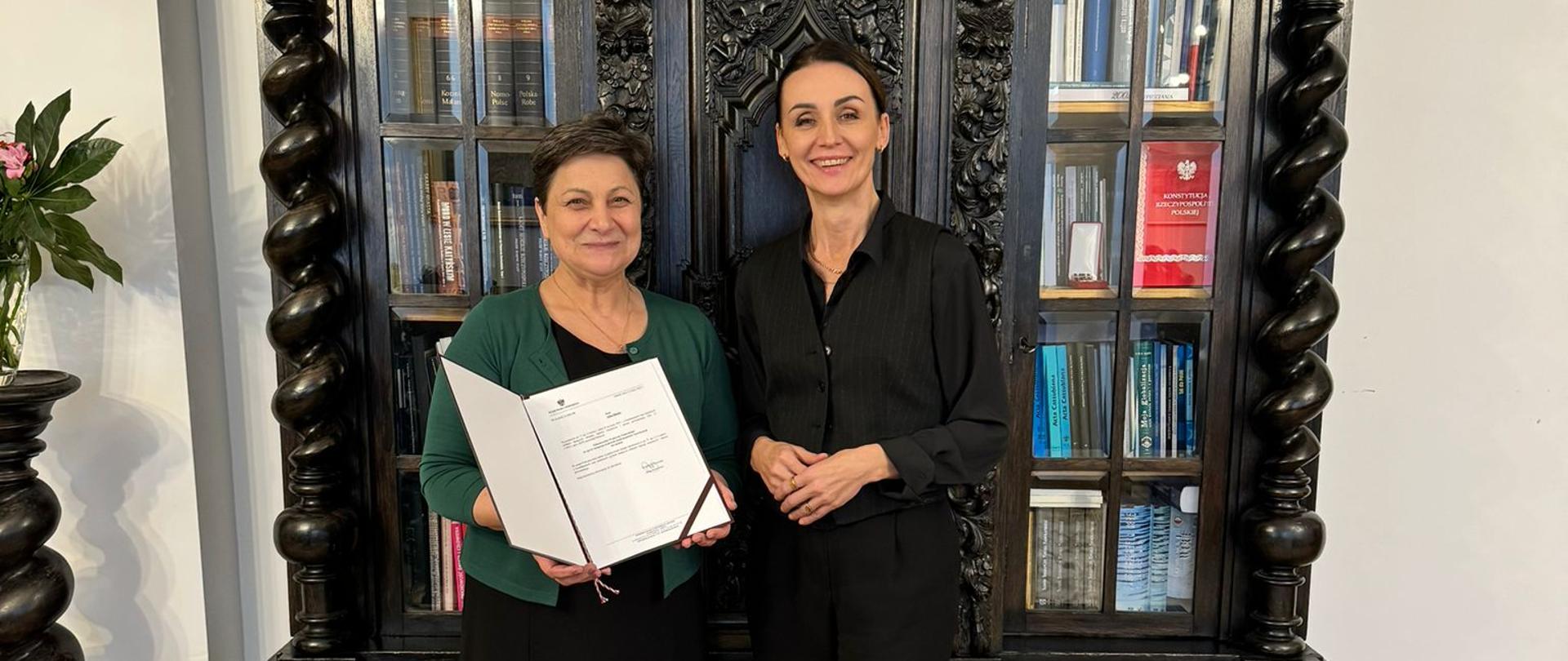 dwie kobiety (wojewoda i pełnomocniczka) stoją w gabinecie na tle zabytkowej biblioteczki