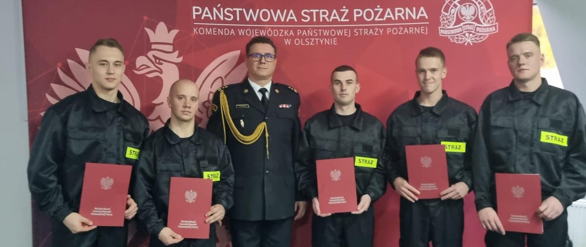 Na tle czerwonego baneru z napisem Państwowa Straż Pożarna stoją strażacy w ubraniu koszarowym w towarzystwie Komendanta Miejskiego Państwowej Straży Pożarnej w Elblągu.