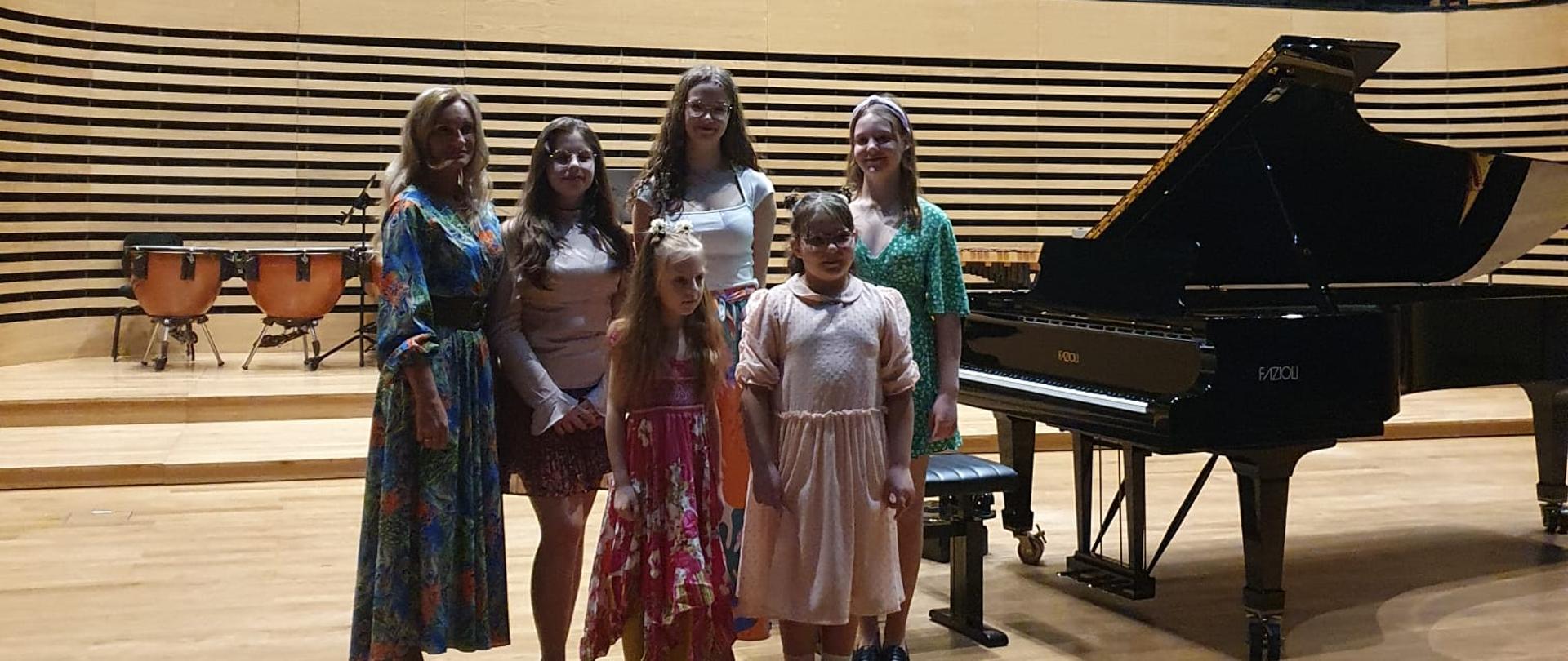 Pięć dziewczynek i kobieta stoją na estradzie sali koncertowej przy fortepianie.