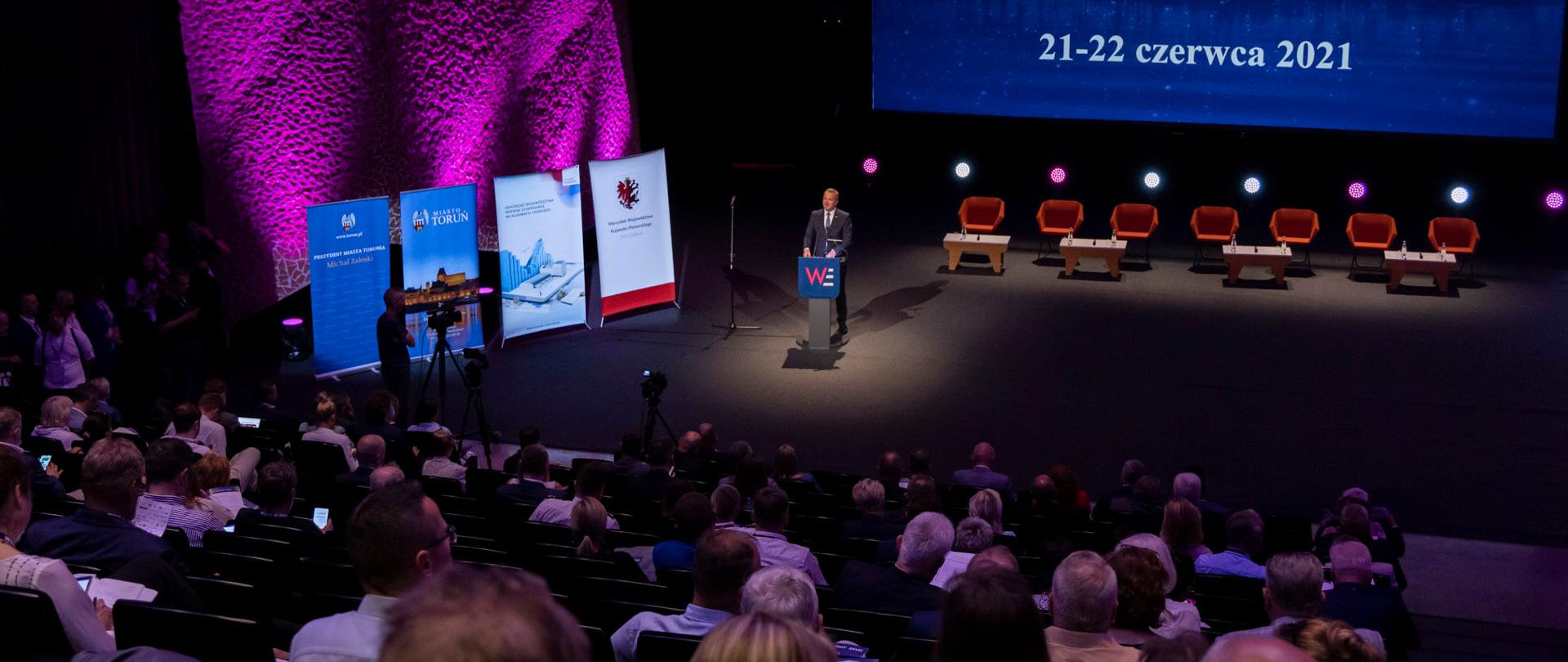 Na zdjęciu widzimy mównicę przy której stoi Wojewoda Kujawsko-Pomorski Mikołaj Bogdanowicz. Przed nim widownia pełna ludzi.