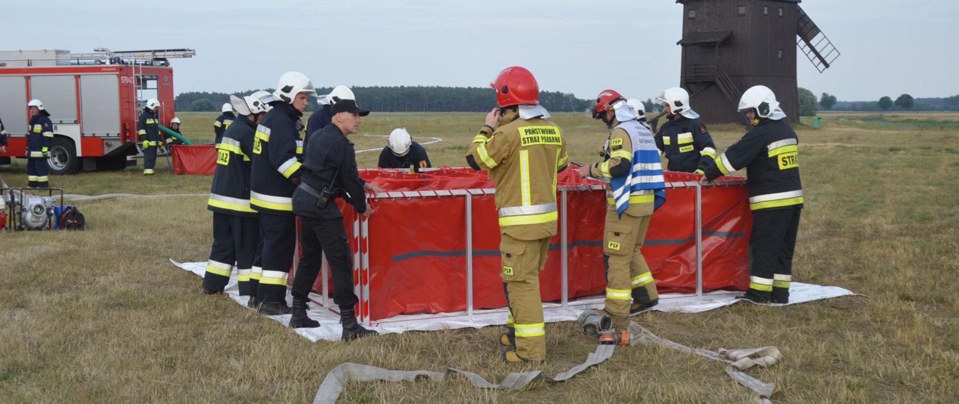 Ćwiczenia "Plutonu Rawicz". Teren nad zbiornikiem retencyjnym w Jutrosinie. Strażacy uczestniczący w ćwiczeniach sprawiają przenośny zbiornik na wodę o pojemności 13 000 litrów. Do zbiornika doprowadzone są linie wężowe. W tle drewniany wiatrak oraz wóz bojowy.