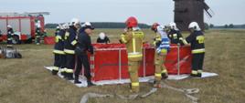 Ćwiczenia "Plutonu Rawicz". Teren nad zbiornikiem retencyjnym w Jutrosinie. Strażacy uczestniczący w ćwiczeniach sprawiają przenośny zbiornik na wodę o pojemności 13 000 litrów. Do zbiornika doprowadzone są linie wężowe. W tle drewniany wiatrak oraz wóz bojowy.