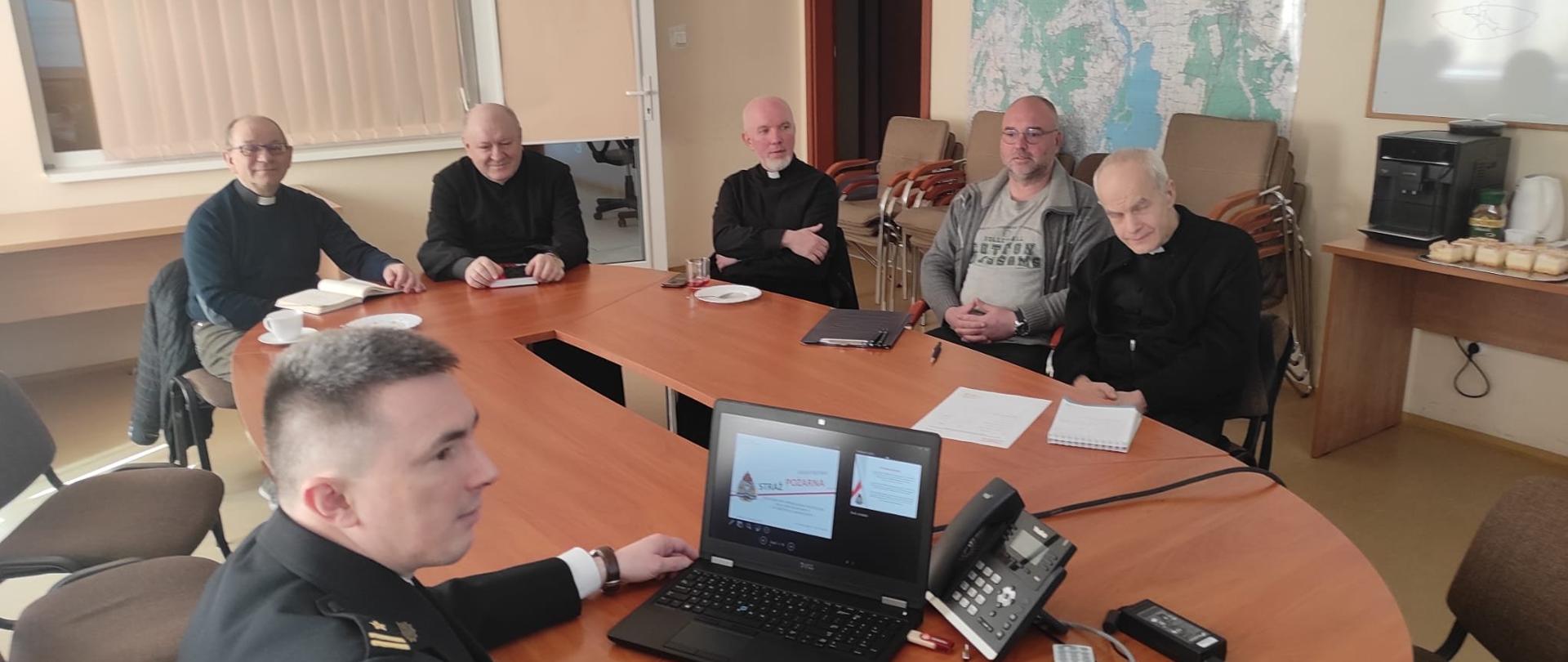 Na zdjęciu sala z owalnym stołem, przy którym siedzi sześciu mężczyzn. Na pierwszym planie widoczny prawy profil twarzy strażaka w mundurze obsługującego przenośny komputer z widoczną prezentacją na ekranie. Za komputerem stojący telefon stacjonarny. Po drugiej stronie stołu na obwodzie widać czterech księży w czarnych sutannach z koloratką oraz mężczyznę w okularach w szarej bluzie. W tle zasłonięte beżową żaluzją okno, mapa, stół z ekspresem do kawy i biała suchościeralna tablica.