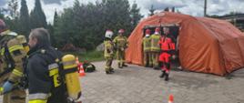 Na zdjęciu widoczni są strażacy zabezpieczeni w aparaty ochrony dróg oddechowych oraz namiot, w którym znajdują się ewakuowane osoby.