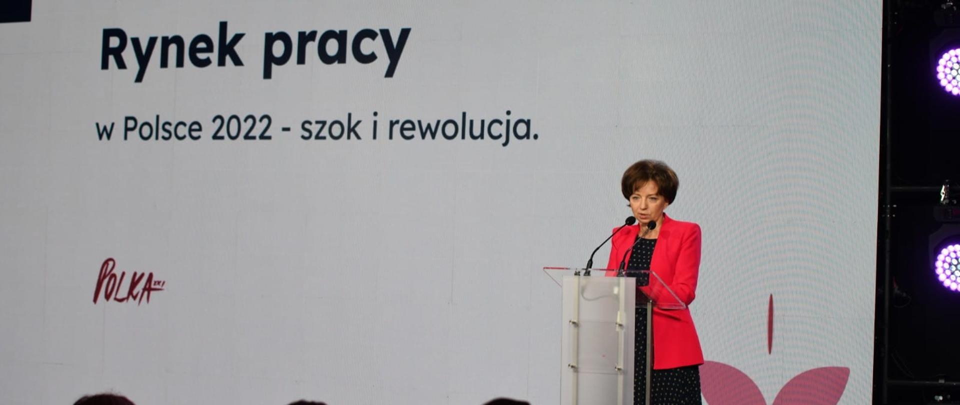 Na tle białego baneru z napisem Rynek pracy w Polsce 2022 - szok i rewolucja, przy mównicy stoi minister Maląg. Przed nią widać zarysy głów słuchaczy.
