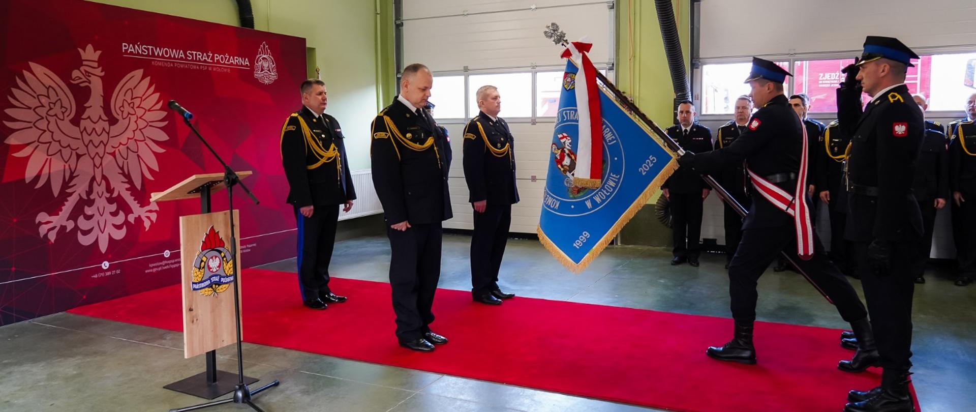 To zdjęcie dokumentuje uroczysty moment pożegnania ze sztandarem st. bryg. Jarosława Nadwodnego, odchodzącego na emeryturę Komendanta Powiatowego PSP w Wołowie. Ceremonia odbywa się z zachowaniem pełnego ceremoniału strażackiego. Na jaskrawym czerwonym dywanie stoją trzej oficerowie w mundurach galowych. Środkowy z nich (odchodzący komendant) stoi ze spuszczoną głową w geście szacunku i skupienia. Po prawej stronie widać poczet sztandarowy. Funkcjonariusz pochyla niebieski sztandar Komendy Powiatowej w Wołowie w kierunku komendanta. Sztandar zdobiony jest złotymi frędzlami oraz biało-czerwoną wstęgą. Obok sztandaru stoi strażak w postawie zasadniczej, który salutuje, oddając honor. Po lewej stronie znajduje się bordowa ścianka z logotypami Państwowej Straży Pożarnej oraz dużym białym Godłem Polski. Obok ścianki ustawiona jest drewniana mównica z mikrofonem, na której widnieje herb formacji. W tle widoczny jest pododdział strażaków stojących wzdłuż białych bram garażowych, co podkreśla wspólnotowy charakter pożegnania. Uroczystość ma miejsce w hali garażowej jednostki, co jest tradycyjnym miejscem dla tak ważnych wydarzeń w strażackiej służbie.
