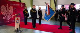 To zdjęcie dokumentuje uroczysty moment pożegnania ze sztandarem st. bryg. Jarosława Nadwodnego, odchodzącego na emeryturę Komendanta Powiatowego PSP w Wołowie. Ceremonia odbywa się z zachowaniem pełnego ceremoniału strażackiego. Na jaskrawym czerwonym dywanie stoją trzej oficerowie w mundurach galowych. Środkowy z nich (odchodzący komendant) stoi ze spuszczoną głową w geście szacunku i skupienia. Po prawej stronie widać poczet sztandarowy. Funkcjonariusz pochyla niebieski sztandar Komendy Powiatowej w Wołowie w kierunku komendanta. Sztandar zdobiony jest złotymi frędzlami oraz biało-czerwoną wstęgą. Obok sztandaru stoi strażak w postawie zasadniczej, który salutuje, oddając honor. Po lewej stronie znajduje się bordowa ścianka z logotypami Państwowej Straży Pożarnej oraz dużym białym Godłem Polski. Obok ścianki ustawiona jest drewniana mównica z mikrofonem, na której widnieje herb formacji. W tle widoczny jest pododdział strażaków stojących wzdłuż białych bram garażowych, co podkreśla wspólnotowy charakter pożegnania. Uroczystość ma miejsce w hali garażowej jednostki, co jest tradycyjnym miejscem dla tak ważnych wydarzeń w strażackiej służbie.