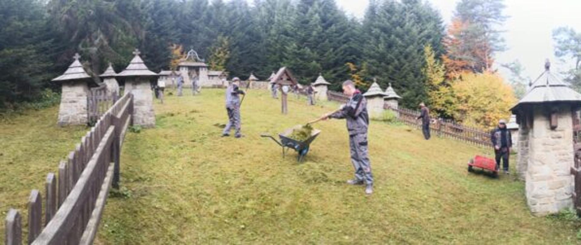 Grupa osób porządkuje teren przy zabytkowym cmentarzu w lesie; widoczne kamienne kapliczki i ogrodzenie.