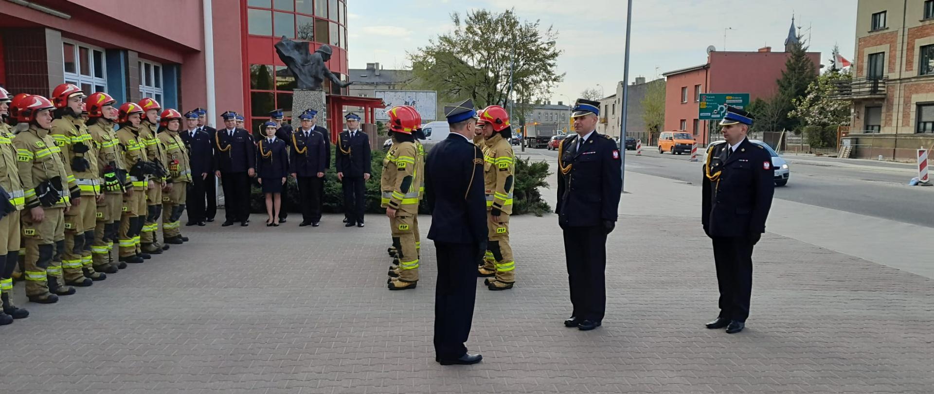 Na zdjęciu funkcjonariusze PSP przed budynkiem JRG. Strażacy w umundurowaniach wyjściowych czarnych oraz piaskowych specjalnych. Na mundurach wyjściowych godło państwowe na rękawie. Osoby posiadają nakrycia głowy. Zdjęcie zrobione w ciągu dnia.