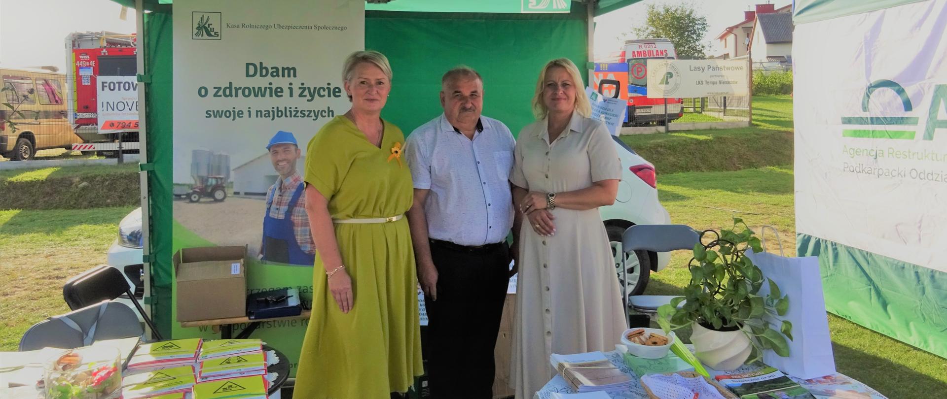 Przy stoisku prewencyjnym Kasy na tle roll-upów stoją dwie kobiety i mężczyzna . Przed nimi znajduje się stół, na którym są wyłożone materiały i ulotki prewencyjne.