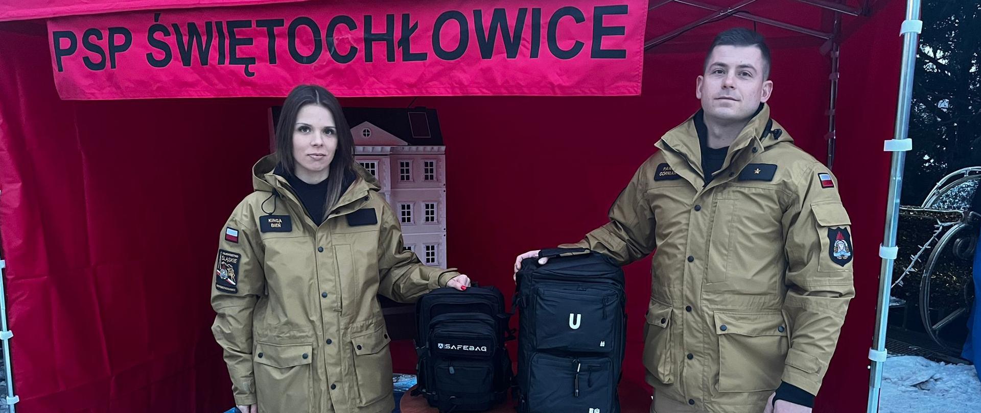 Zdjęcie przedstawia dwoje funkcjonariuszy Państwowej Straży Pożarnej (PSP) ze Świętochłowic, kobietę i mężczyznę, stojących pod czerwonym namiotem promocyjnym. Są oni ubrani w profesjonalne, piaskowe mundury służbowe z widocznymi naszywkami jednostki oraz flagą Polski na ramionach. Sceneria wskazuje na warunki zimowe, ponieważ podłoże pokryte jest warstwą śniegu i lodu.
Para prezentuje dwa czarne plecaki ratunkowe lub ewakuacyjne, które spoczywają na niewielkim, okrągłym stoliku między nimi. W tle, pod zadaszeniem namiotu, widoczny jest model domku, który służy jako pomoc dydaktyczna do edukacji o bezpieczeństwie pożarowym. 
