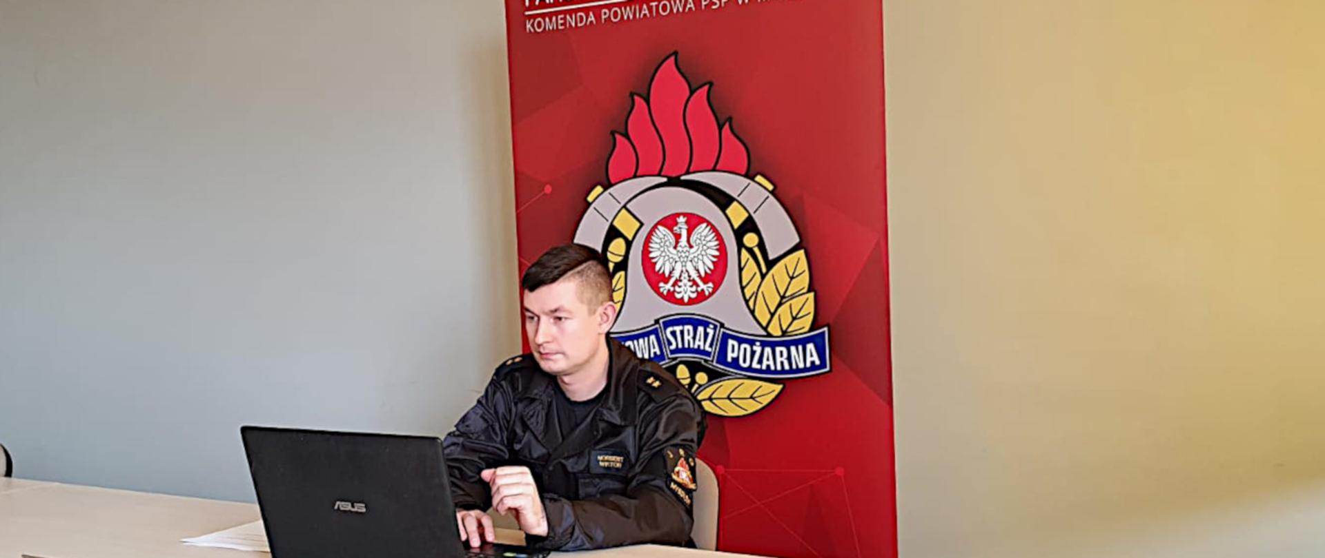 Na zdjęciu widoczna jest sala konferencyjna w komendzie. Przy stole siedzi strażak w ubraniu dowódczo sztabowym. Na stole, przed nim leży laptop. W tle logo PSP i napis Komenda Powiatowa PSP w Myszkowie.