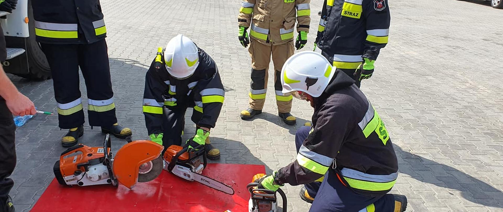 Zdjęcie przedstawia pięciu strażaków w ciemnych ubraniach ochronnych i hełmach podczas ćwiczeń pilarkami i przecinarkami spalinowymi na utwardzonym placu.
