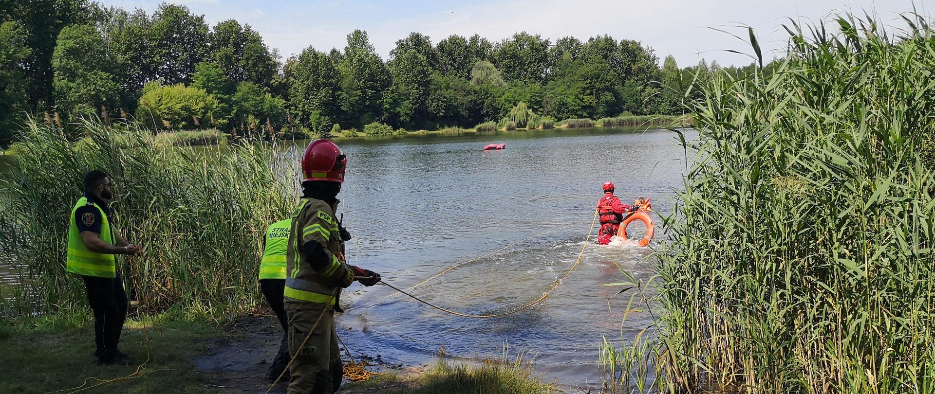 Zdjęcie przedstawia strażaka w wodzie niosącego koło. Drugi strażak asekuruje go linką ratowniczą.