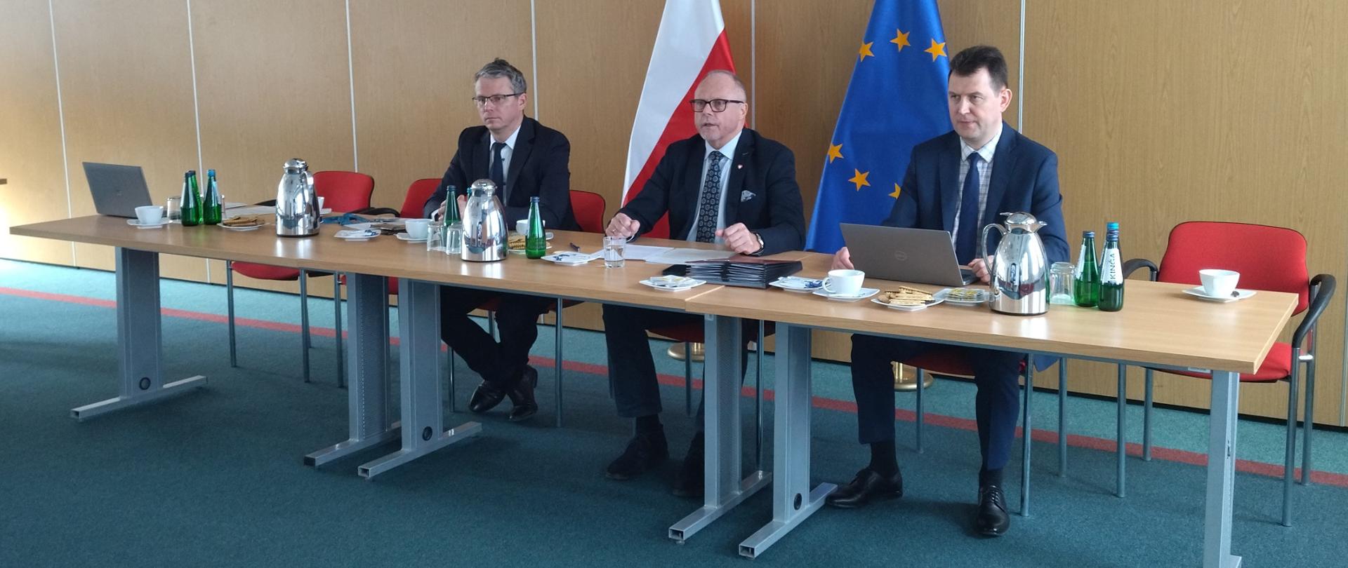 Trzech mężczyzn siedzi przy stole. W środku wiceminister Jacek Protas. Za nimi flagi PL i UE.