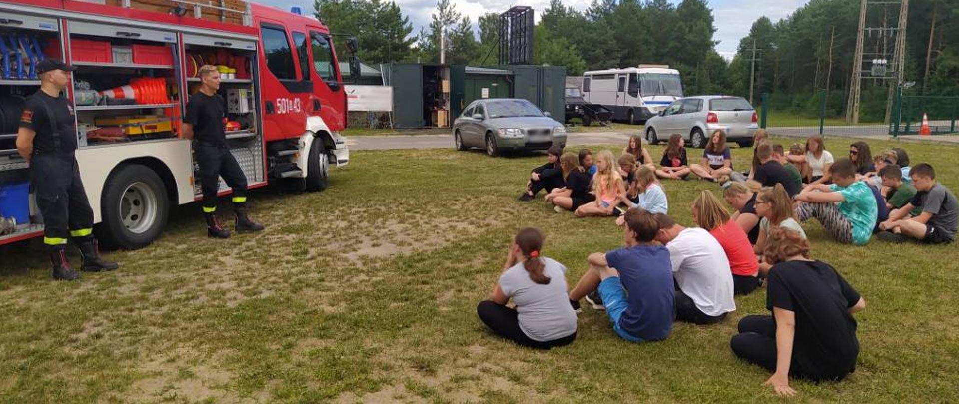 Zdjęcie przedstawia uczestników obozu harcerskiego – młodzież w wieku 11-18 lat, siedzących na trawie podczas spotkania profilaktycznego z funkcjonariuszami Komendy Powiatowej Państwowej Straży Pożarnej w Zambrowie. Prowadzący pogadankę dwaj funkcjonariusze ubrani w krótką czarną koszulkę z logiem PSP oraz spodnie z ubrania koszarowego. Młodzież w ubraniach cywilnych. Za stojącymi frontem do uczestników obozu strażaków stoi średni samochód ratowniczo-gaśniczy z Jednostki Ratowniczo-Gaśniczej w Zambrowie z otwartymi kurtynami w celu prezentacji znajdującego się na pojeździe sprzętu. Zdjęcie wykonano na terenach zielonych Ośrodka szkolenia poligonowego Grom Group w Czerwonym Borze w słoneczny dzień. 
