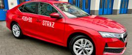 Czerwony samochód marki Skoda. Biały napis Straż oraz boczny numer operacyjny znajdujący się na boku pojazdu. Stoi na kostce pol brukowej a w tle niebieskie bramy garażowe budynku KM PSP w Gdyni.