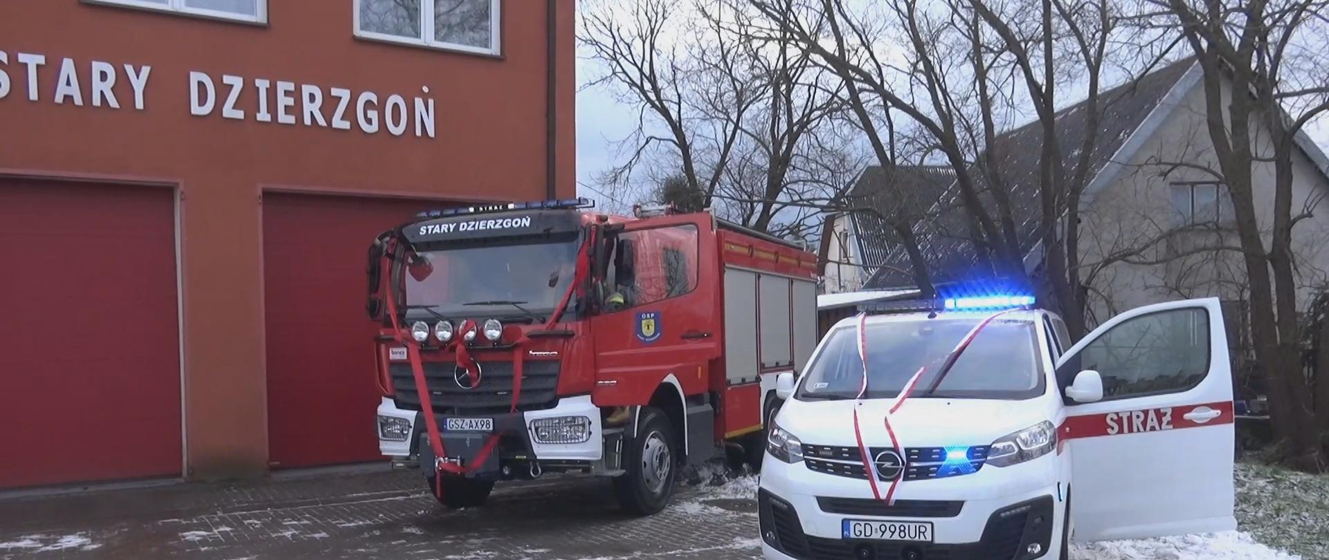 Pora dzienna, kadr przedstawia od lewej: usytuowana remiza OSP z czerwoną elewacją, kolejno pojazd pożarniczy w kolorze czerwonym oraz biały bus z otwartymi drzwiami kierowcy z prawej strony.. Pojazdy mają przewieszone szarfy z przodu.