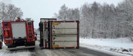 Przewrócona na drodze naczepa samochodu ciężarowego. Obok stoi samochód strażacki. Droga i pobocze zaśnieżone.
