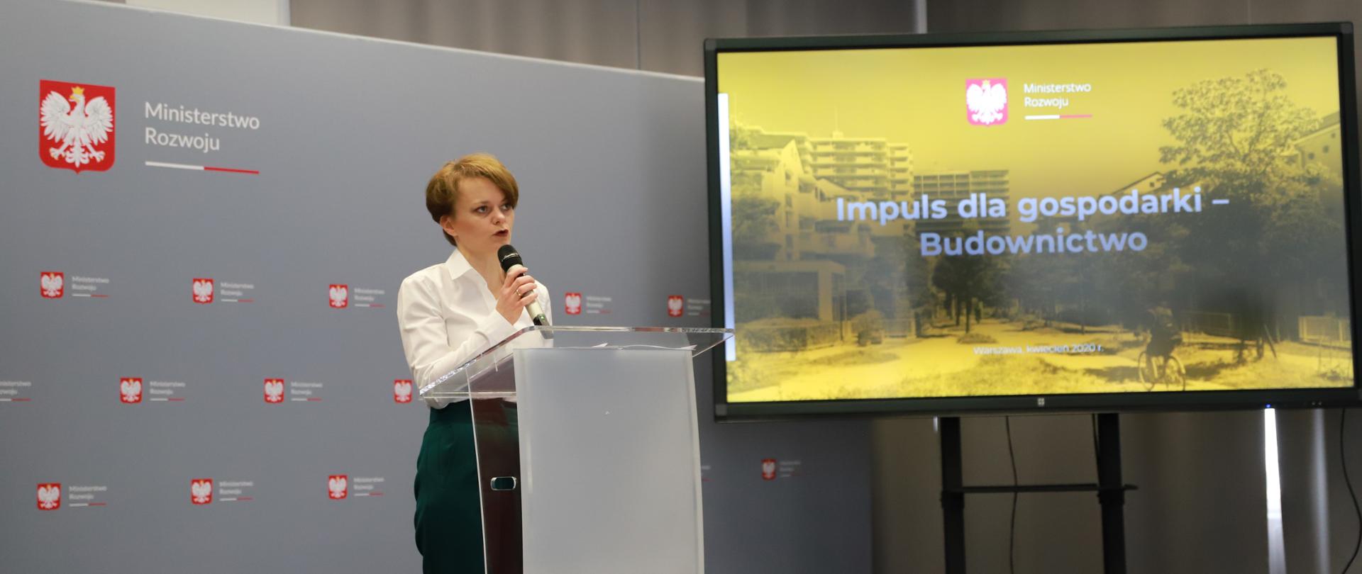 Wicepremier Jadwiga Emilewicz w trakcie przemówienia. Stoi za mównicą, na tle ścianki z logo MR, po prawej stronie ekran, na którym wyświetla się hasło Impuls dla gospodarki - Budownictwo.